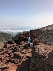Fototapeta premium Man standing on top of a mountain over sea of clouds. Adventurer looking at vast scenic landscape. Backpacker enjoying outdoor exploration and freedom in high altitude on sunny day. Travel and wellnes