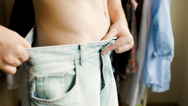 man showing loose jeans after weight loss and body transformation. He gives a thumbs up, representing healthy habits and lifestyle. High quality 4k footage