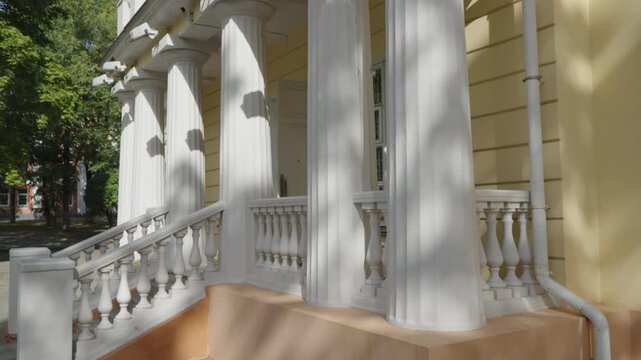 White classical columns and a sculpted balustrade frame the grand entrance porch of a historic yellow mansion on a sunny day.