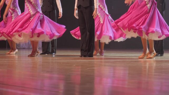 Female ballroom dancers wearing vibrant pink dresses and male partners in black trousers perform a synchronized dance routine on a glossy wooden floor.