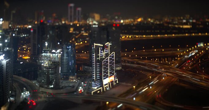 Dubai at night, lit office towers stand beside a wide highway junction, car light trails cross the ramps, construction cranes work nearby, tilt shift miniature look, timelapse