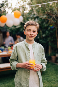 Birthday celebration theme for party event. A young boy holding a glass of orange juice in his hand, standing in a grassy area surrounded by trees and balloons. In the background.