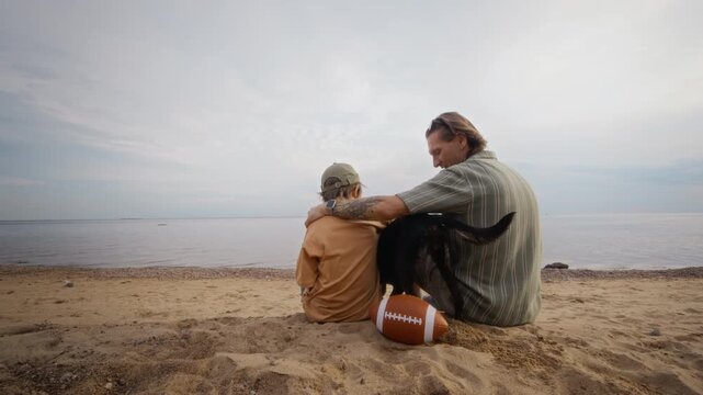 Caucasian father and child son sitting on sandy beach, petting black dog next to American football. Clip suitable for promoting family travel or outdoor leisure activities