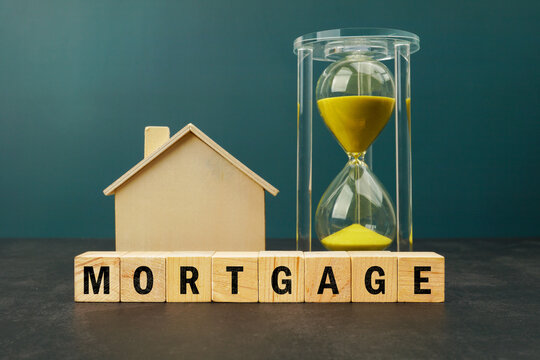 Wooden house model beside an hourglass and wooden blocks spelling MORTGAGE. Concept of home financing, property ownership, mortgage planning and long term financial commitment.