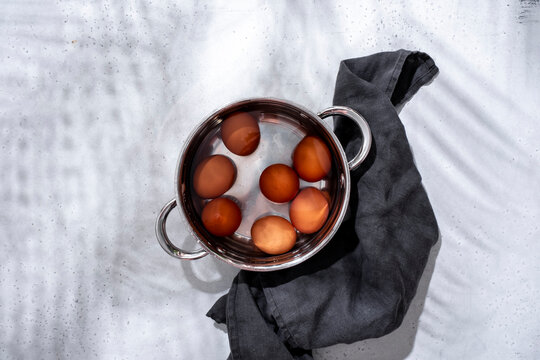 Brown chicken eggs submerged in water in pot, accompanied by dark grey linen towel on light grey background in hard light.