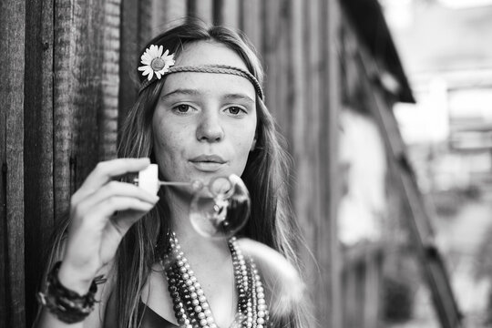 Black and white portrait of a teenage girl blowing soap bubbles while wearing a flower headband and layered bead necklaces outdoors