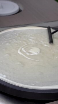 Close-up shot of a gloved hand using a T-shaped wooden spreader to smooth and flatten thin liquid batter across a hot round crepe griddle.