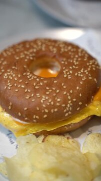 Close-up shot of a gourmet breakfast sandwich made with a toasted sesame seed bagel, folded fluffy egg, and melted cheese, served on patterned paper with crispy potato chips on a plate.