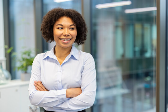 Close-up photo of a young African American businesswoman standing in the office, arms crossed on her chest and looking to the side with a smile