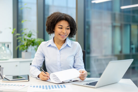 Smiling young African American woman working in office with paperwork and looking at laptop screen