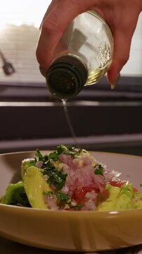 Vertical video. Woman Pours Cooking Oil into Bowl of Avocado, Tomato, Onion and Cilantro, Preparing Fresh Homemade Guacamole in Sunlit Kitchen