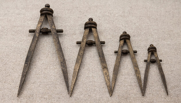Four vintage metal compasses of varying sizes arranged in a row on a textured beige fabric background, for technical work