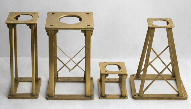 Four different sizes of brass metal stands with square bases and cross-braced supports standing on a plain white background surface