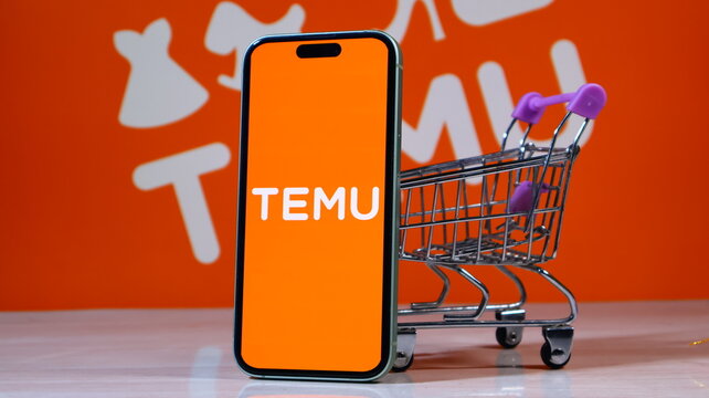 Smartphone displaying Temu logo next to miniature shopping cart on orange background.