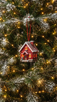 Small red house ornament hanging on a glowing green christmas tree with warm fairy lights