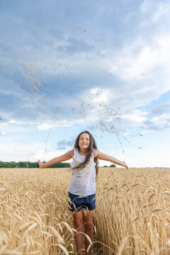 Happy little girl throwing wheat spikelets in air in golden field under blue sky