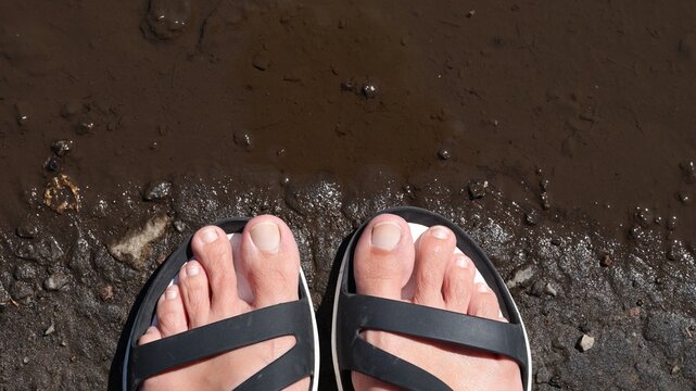 A top view of bare human feet in black sandals standing on muddy ground next to a brown puddle.