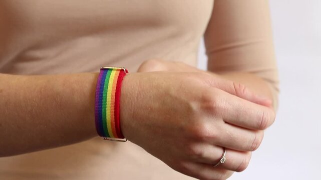 Woman fastens rainbow bracelet on wrist. Close-up of a woman's hands wearing a rainbow colored thong (LGBTQ+ pride) against a white wall. LGBT pride concept: a person with a rainbow accessory