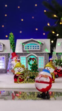 A quirky Santa Claus and a yellow car appear in front of a decorated Christmas village