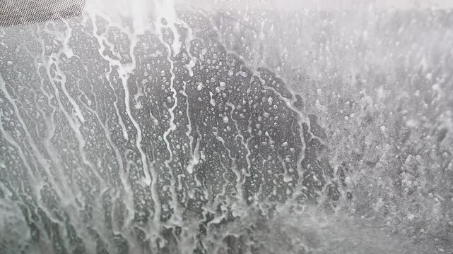 White soap foam sliding down a vehicle's windshield during an automatic car wash, with water rinsing off the dirt and revealing the clean, wet glass underneath in a close-up view
