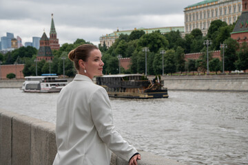 Fototapeta premium Woman glances back beside Moscow River on Sofiyskaya Embankment with Kremlin skyline in background. Urban travel, leisure, architecture, riverfront, tourism, modern lifestyle, freedom