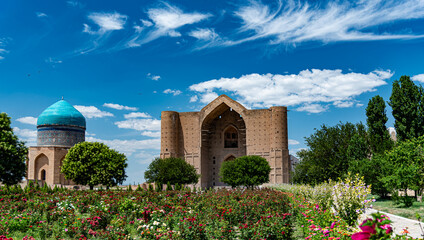 Naklejka premium The majestic Mausoleum of Khoja Ahmed Yasawi made of sandstone with blue domes in Turkestan. Ancient Central Asian architecture against a blue sky with clouds and green trees in the foreground.