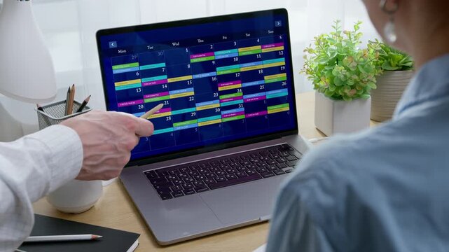 Colleague pointing pen at laptop screen displaying monthly calendar with dates for project planning. Woman reviewing schedule during time management and workplace organization.