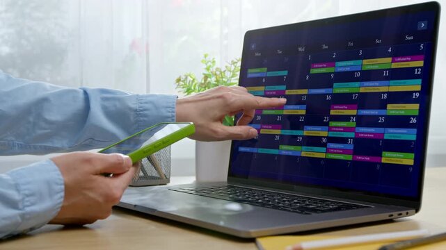 Hands of woman holding smartphone near laptop screen displaying monthly calendar for project planning. Person structuring daily workflow during time management and schedule organization.