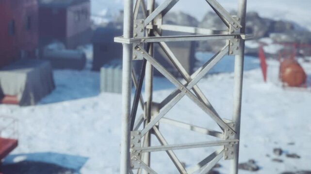 Workers are setting up a tower in a cold area covered in snow. Nearby buildings are visible, and equipment is arranged around the construction site. The scene shows a busy atmosphere.