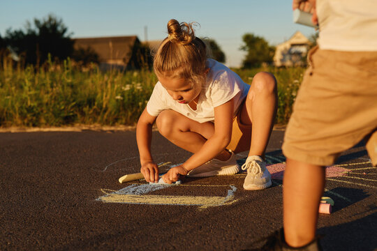 Two children draw pictures with chalk on the asphalt. It is evening and the sun is setting in a neighborhood with green fields