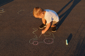 A child is kneeling on the ground using colored chalk to draw a vehicle on the pavement. The sun casts long shadows as the day ends © sementsova321