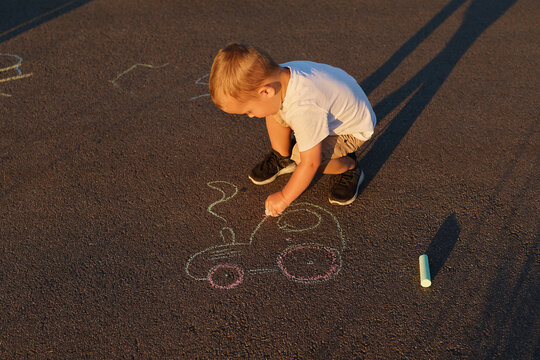 A child is kneeling on the ground using colored chalk to draw a vehicle on the pavement. The sun casts long shadows as the day ends