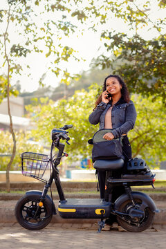 Young Black woman on the phone beside her electric bicycle, preparing for her daily commute to work in a sustainable, eco-friendly lifestyle.