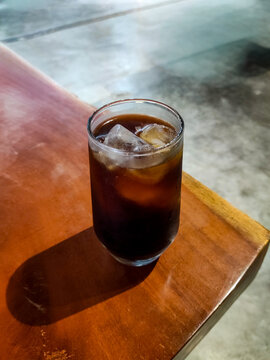 Iced Americano served in a tall glass at the edge of a wooden caf&eacute; table, featuring rich dark coffee, melting ice cubes, and a relaxed atmosphere for casual coffee moments.