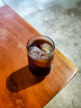 Iced Americano served in a tall glass at the edge of a wooden caf&eacute; table, featuring rich dark coffee, melting ice cubes, and a relaxed atmosphere for casual coffee moments.