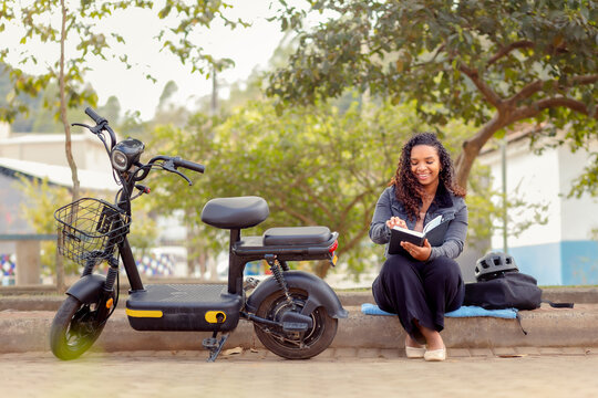 A young Black Brazilian woman takes a break from her daily commute on an electric bicycle to read outdoors in a quiet urban neighborhood.