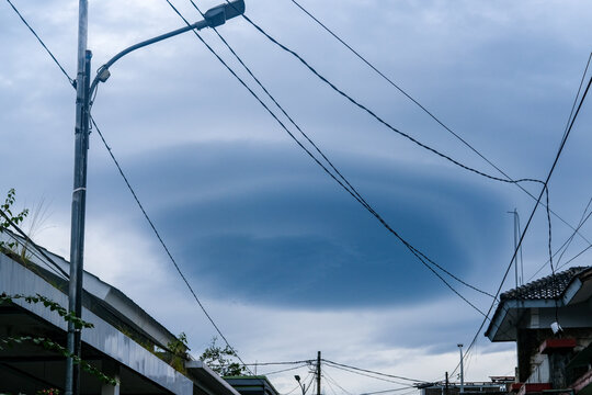 Dramatic circular storm cloud formation over a residential neighborhood, framed by rooftops, utility poles, and overhead wires beneath a moody gray sky before rainfall.