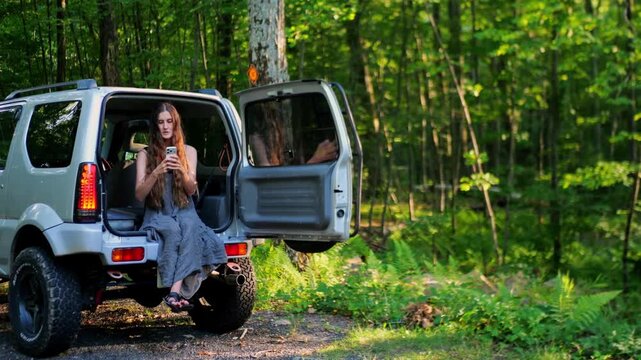 Caucasian woman sitting in open SUV trunk using a smartphone in a green forest, representing outdoor travel, mobile connection and modern adventure lifestyle.