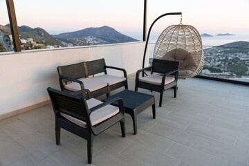 Obraz premium Modern outdoor patio seating and a hanging rattan chair on a tiled terrace overlooking a coastal town and sea at sunset. Comfortable cushions, panoramic mountain and ocean views create a relaxing mood