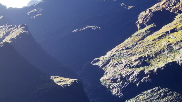 Rocky mountain formation is visible under bright sunlight. Shadows create contours on the surface. Green patches of grass are scattered among the rocks. The scene shows a natural area.