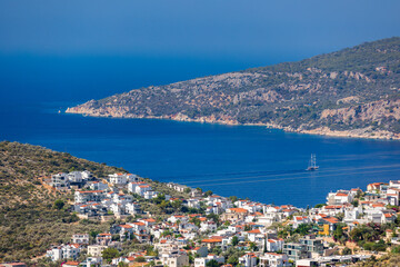 A sunlit coastal town Kalkan cascades down a hillside toward a deep blue bay, where a classic sailboat crosses calm waters beneath rugged Mediterranean hills and a clear summer sky. Turkey © evannovostro