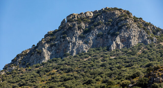 Sunlit rocky mountain peak rises above dense green shrubland on a Mediterranean hillside, creating a rugged natural landscape mood under clear blue sky, evoking solitude and adventure. Kalkan, Turkey