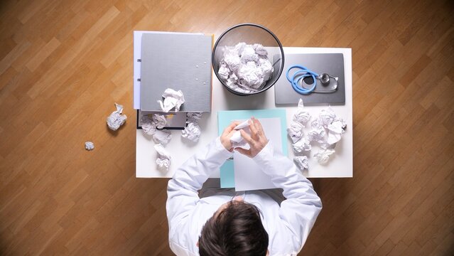 Medical professional experiencing burnout and work pressure, sitting at a desk and crumpling frustrated documents into a trash can full of crumpled paper, surrounded by office mess