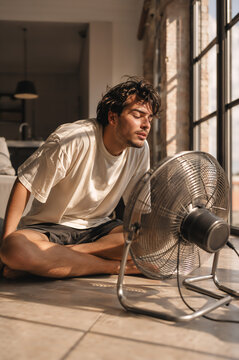 Man sitting on floor in front of electric fan to cool off during hot summer day. Tired male feeling heat exhaustion relief in sunlit room. Indoor cooling appliance in warm weather.
