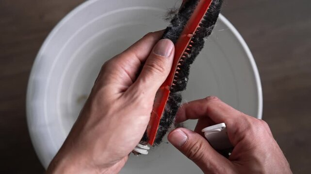 Top view of male hands using small tool to clean tangled hair and dust from vacuum cleaner brush. Close-up of household maintenance, cleaning appliance parts over white bucket for better performance.