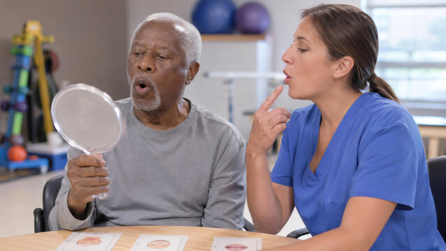 Speech therapist instructing senior man during rehabilitation session. Patient practicing facial muscle exercise with mirror in clinic. Professional health care and recovery assistance.
