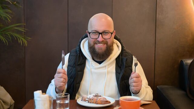 Bald man with glasses eagerly holds knife and fork, gazing at scrumptious dessert plate in charming cafe filled with soft lighting. Moment captures anticipation and delight in tasty experience.