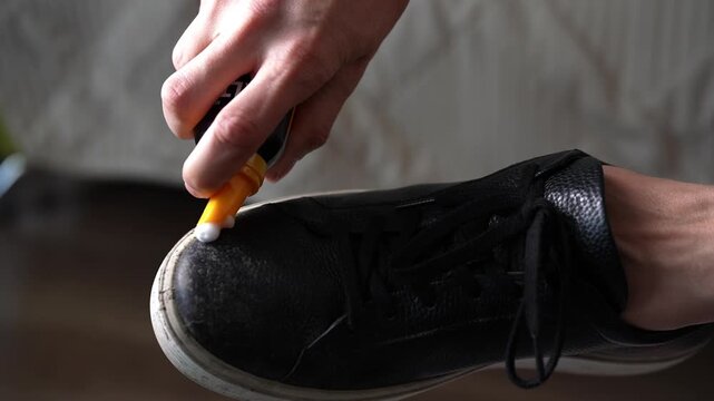 Close-up hands of unrecognizable person applying special white cleaning foam from bottle with sponge applicator to sole of black leather sneaker. Concept of detailed footwear care routine.