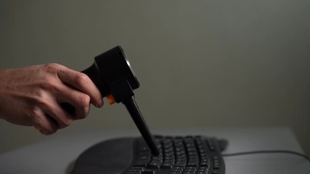 Close-up of hand holding electric air duster blowing dust from black computer keyboard. Concept of cleaning electronics, computer maintenance, hygiene in workspace. Shooting in slow motion.