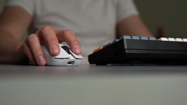 Close-up of person hand using white wireless mouse. Fingers click and scroll. Another hand appears to type on mechanical keyboard. Modern office, gaming setup for work, play. Shooting in slow motion.